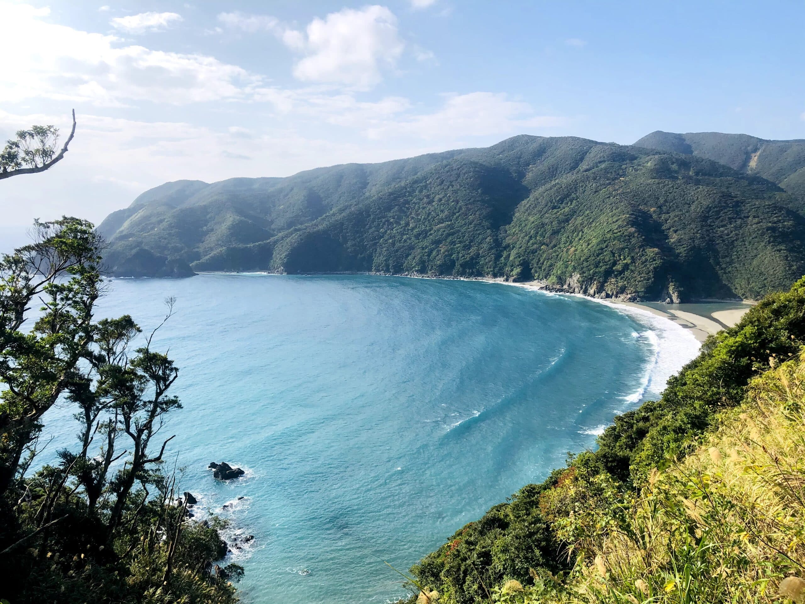 A panoramic view of a lush green coastline and a curved turquoise bay, seen from a high vantage point on Katoku beach