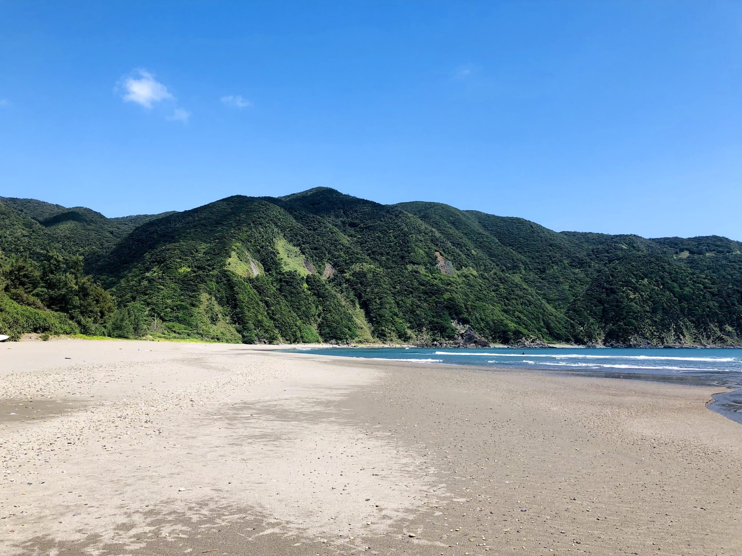 A wide sandy beach backed by forested mountains under a clear blue sky in Katoku beach