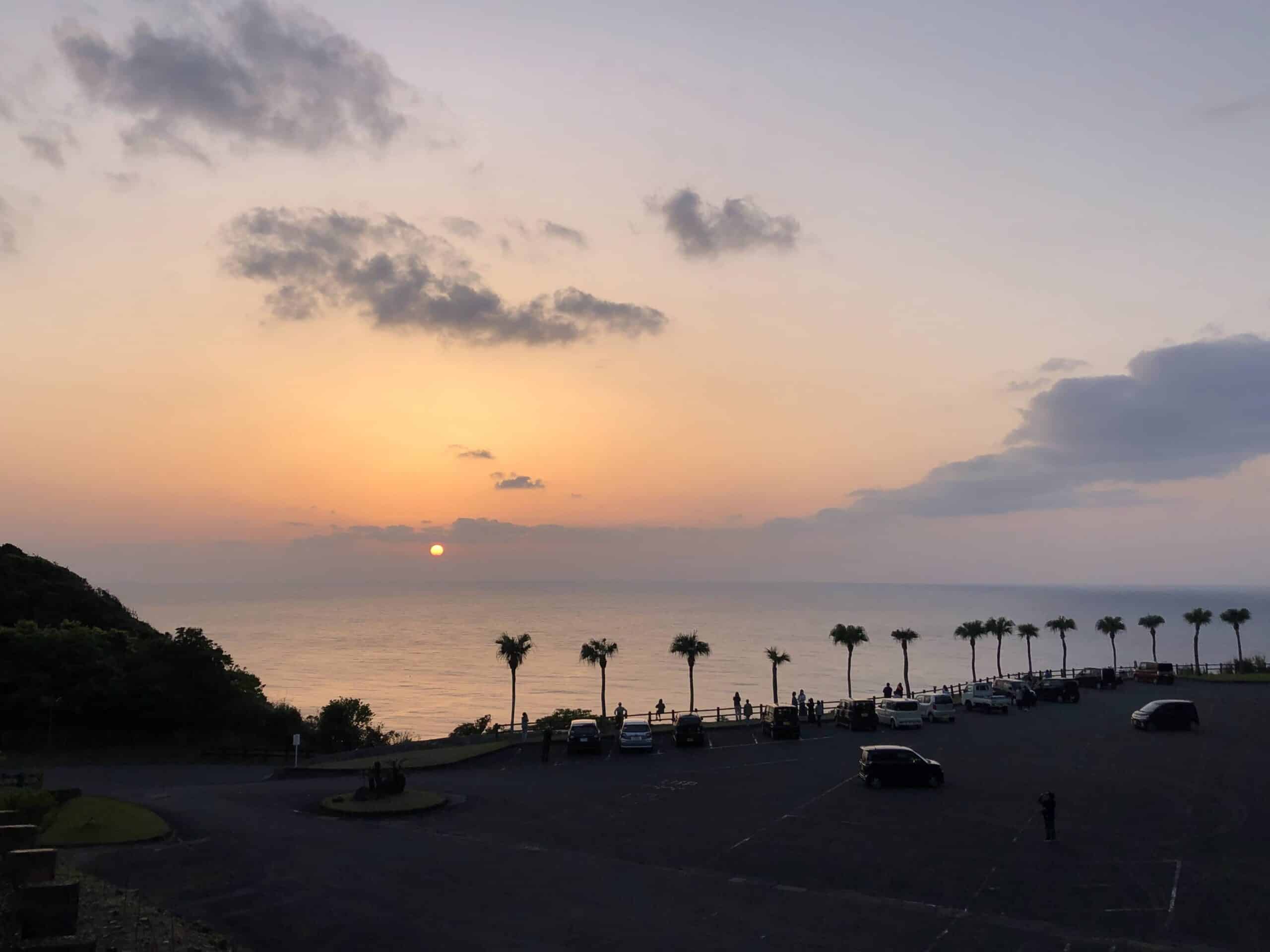 Sunset over the ocean seen from a coastal viewpoint with palm trees, parked cars, and silhouetted visitors on Amami Island.