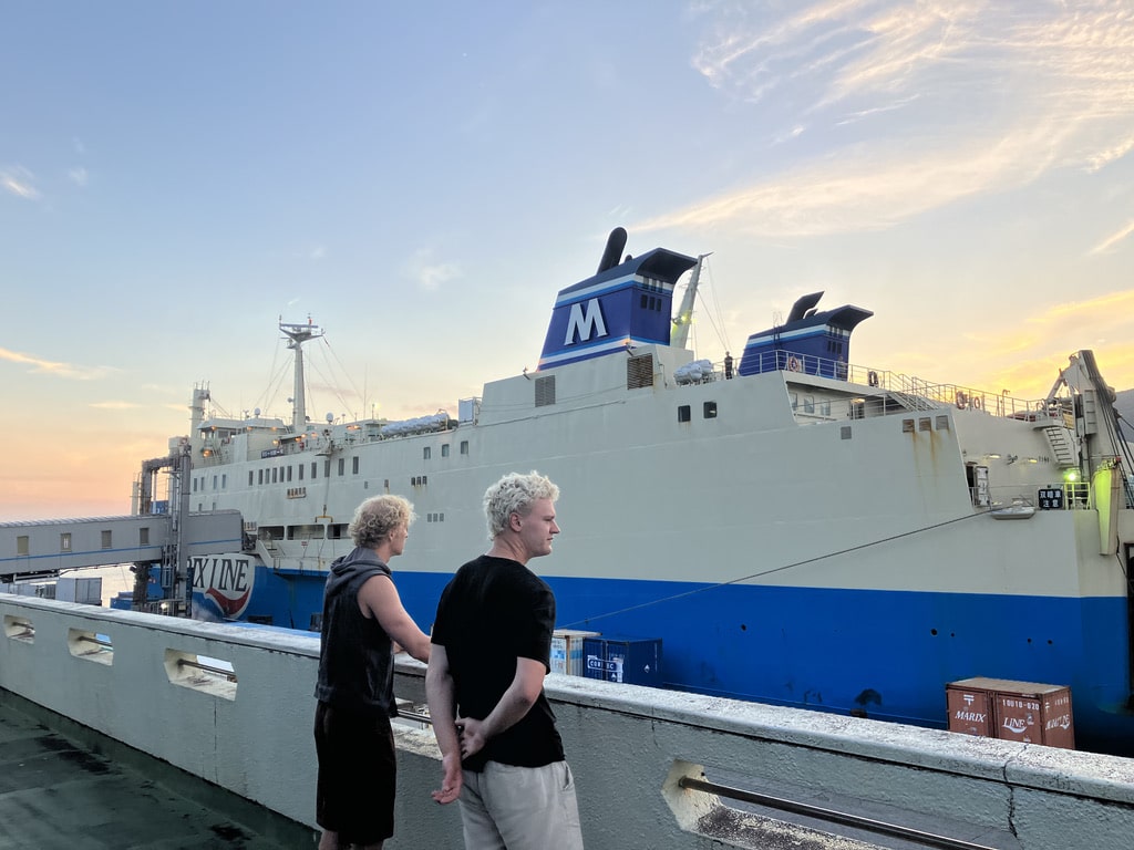 Two foreign travelers standing on the deck, looking at the ferry docked at Naze Port in Amami at sunrise.