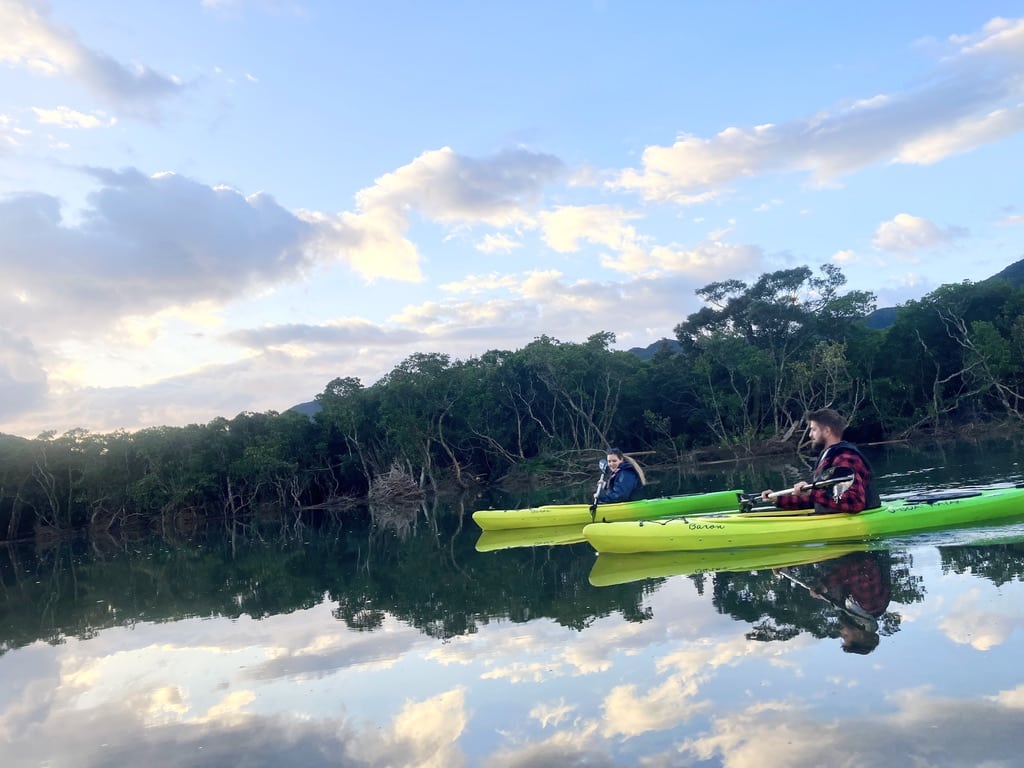 Two travelers kayaking in the calm mangrove river of Amami Oshima at sunrise, reflecting trees and sky.