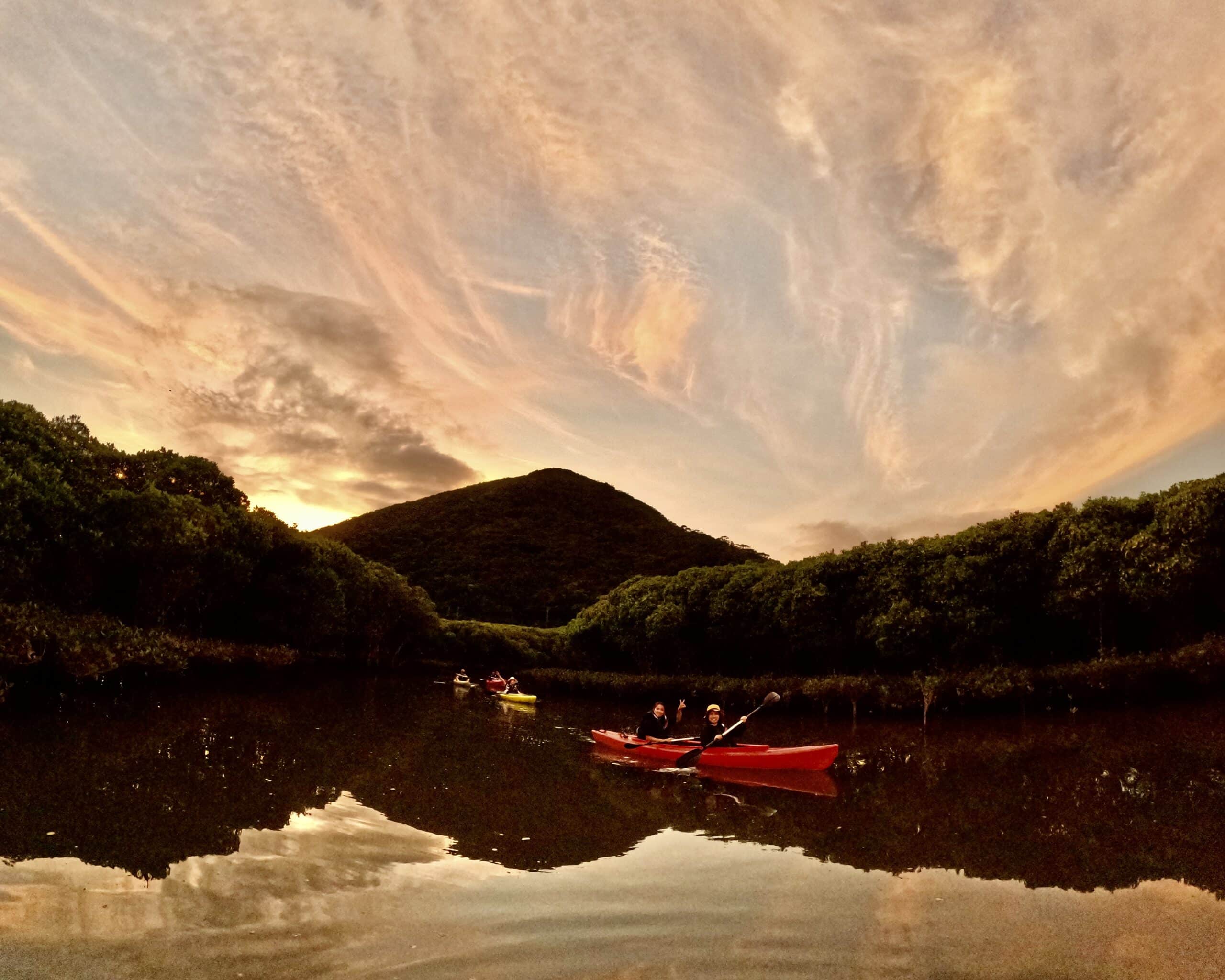 Amami Oshima sunset mangrove kayak tour – paddlers gliding on calm waters with vivid orange and pink sky reflecting on the river