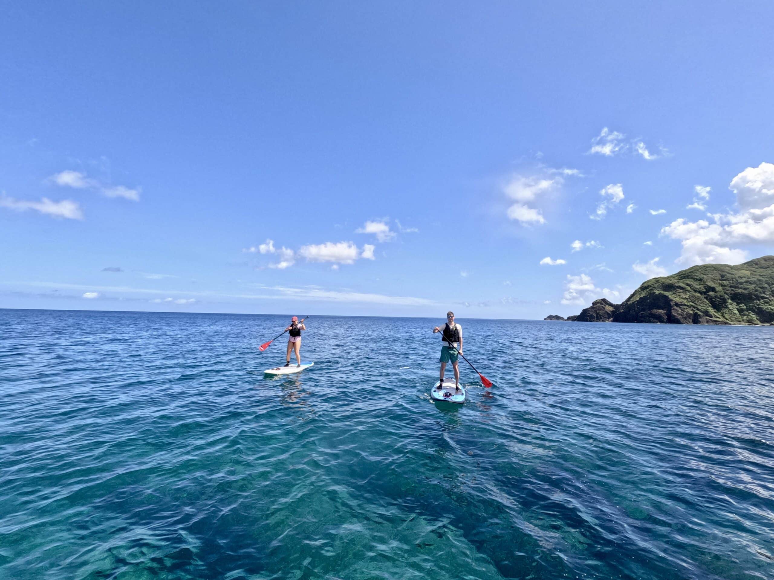 Two people paddleboarding on the open sea with Amami Oshima’s coastline in the background.