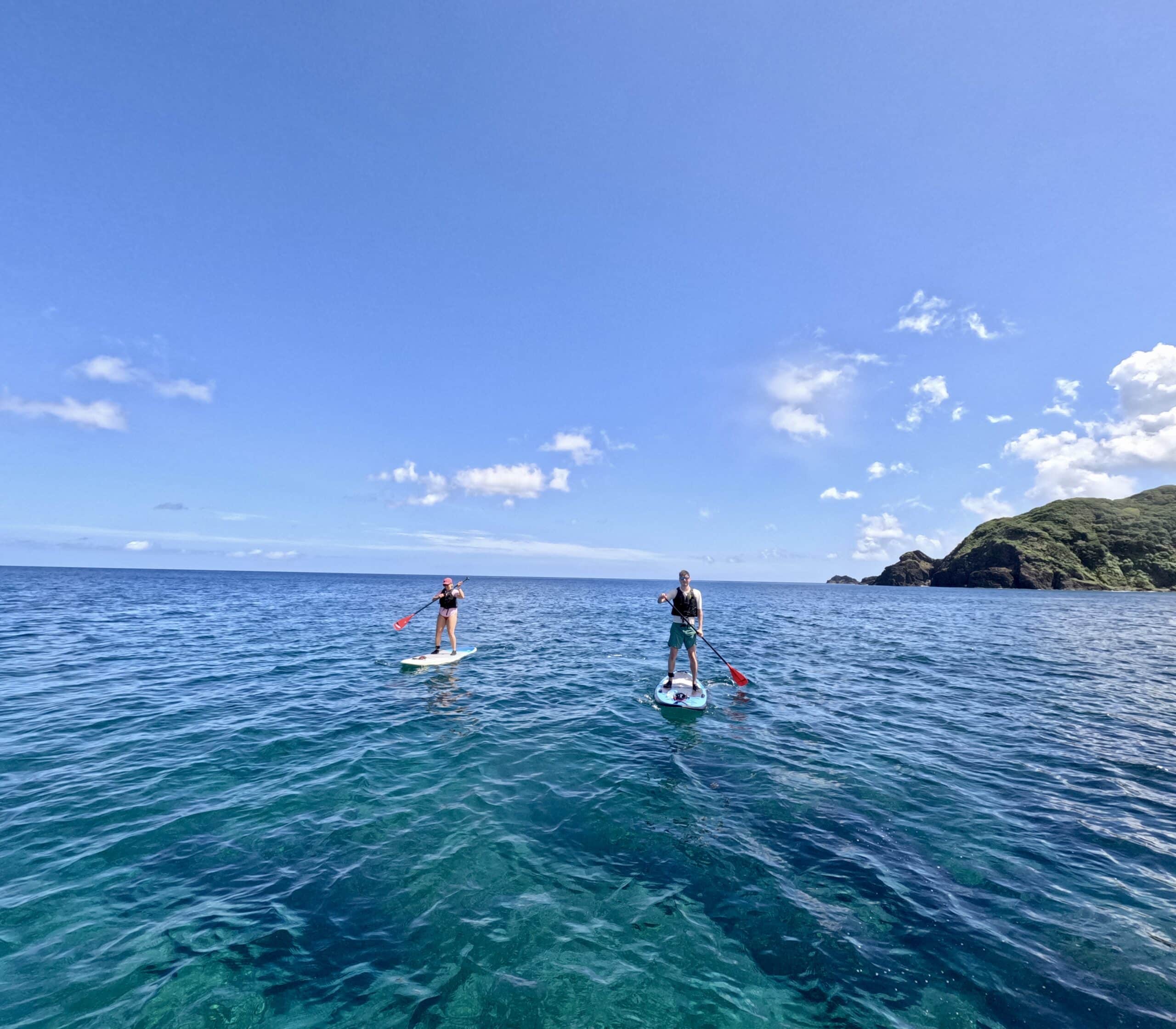 Two people paddleboarding on the open sea with Amami Oshima’s coastline in the background.