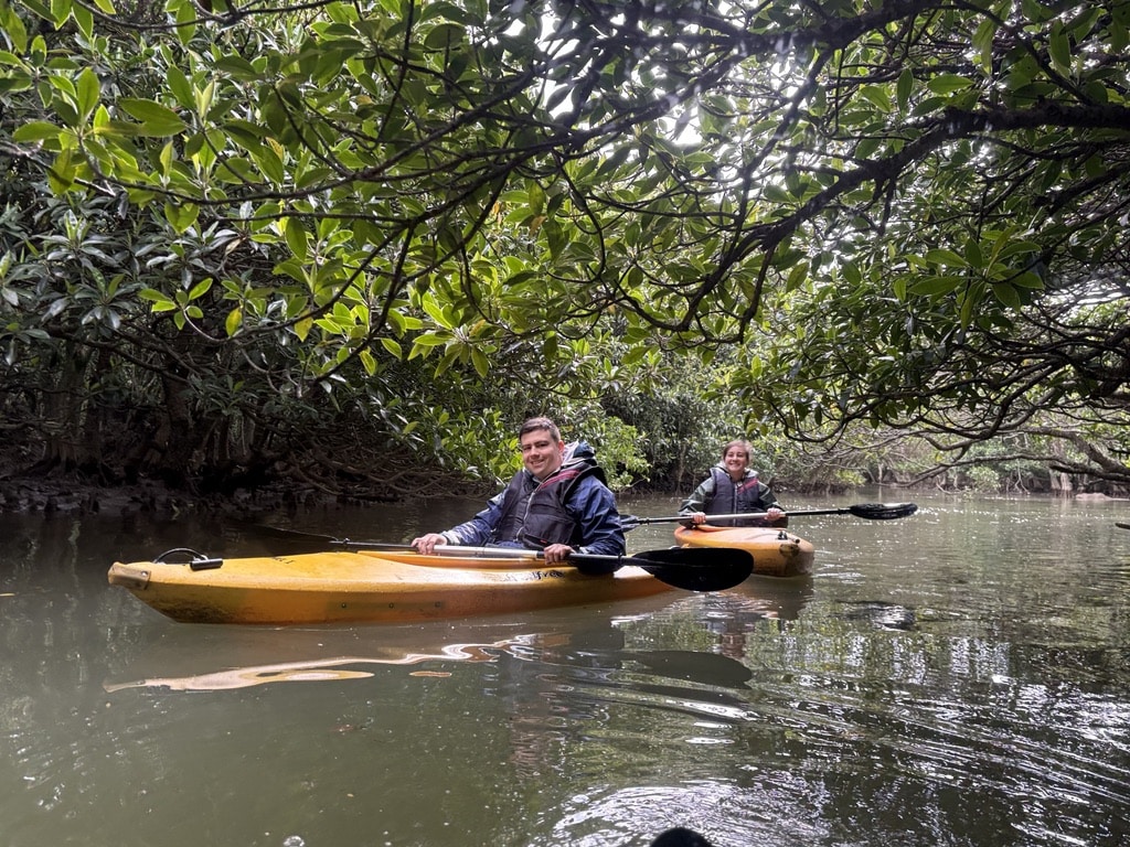 Two guests kayaking through a mangrove tunnel in Amami Oshima during a calm rainy day, surrounded by lush green branches.