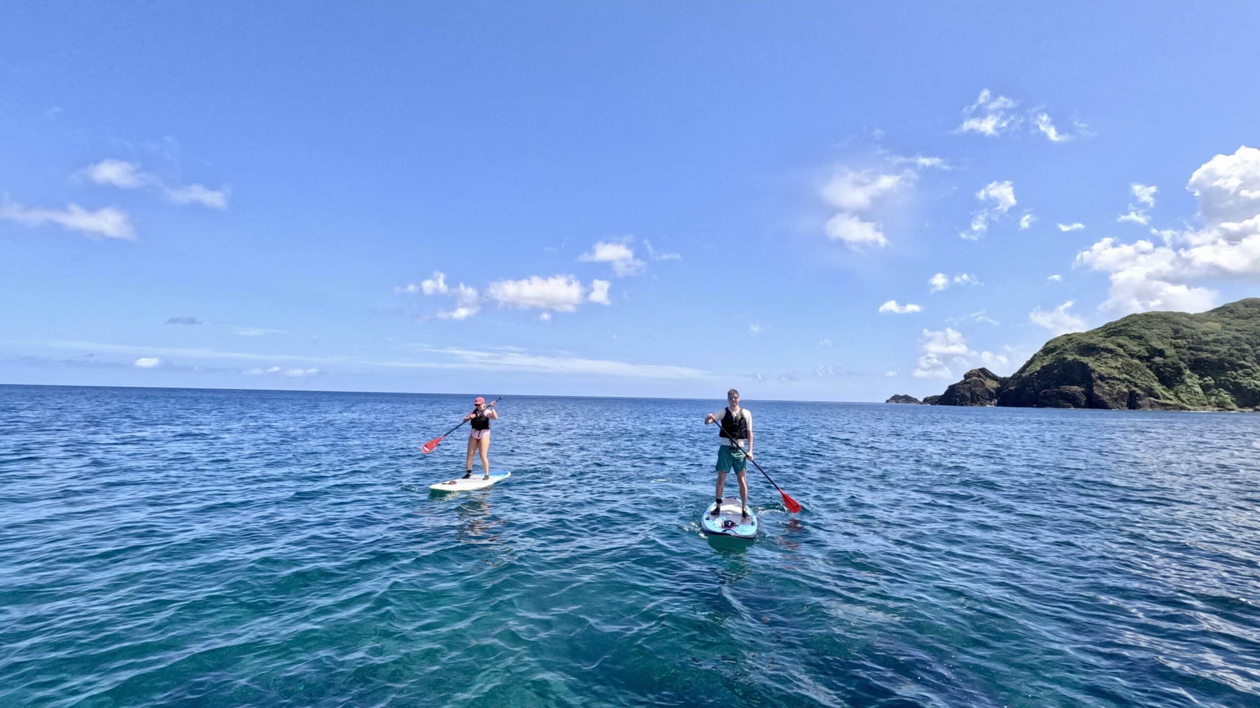 Two people paddleboarding on the open sea with Amami Oshima’s coastline in the background.